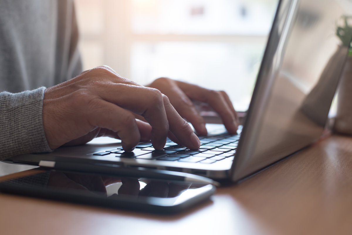 Young man or freelancer using laptop computer, searching web with smart phone on wooden table and morning light, browsing internet information, working from home office concept, close up